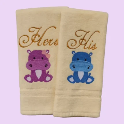 Cream towels with “Hers” and “His” embroidery and cute pink and blue hippos on pastel background