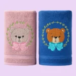 Pink and blue towels with cute bear faces and bows embroidered inside leaf wreaths on pastel background